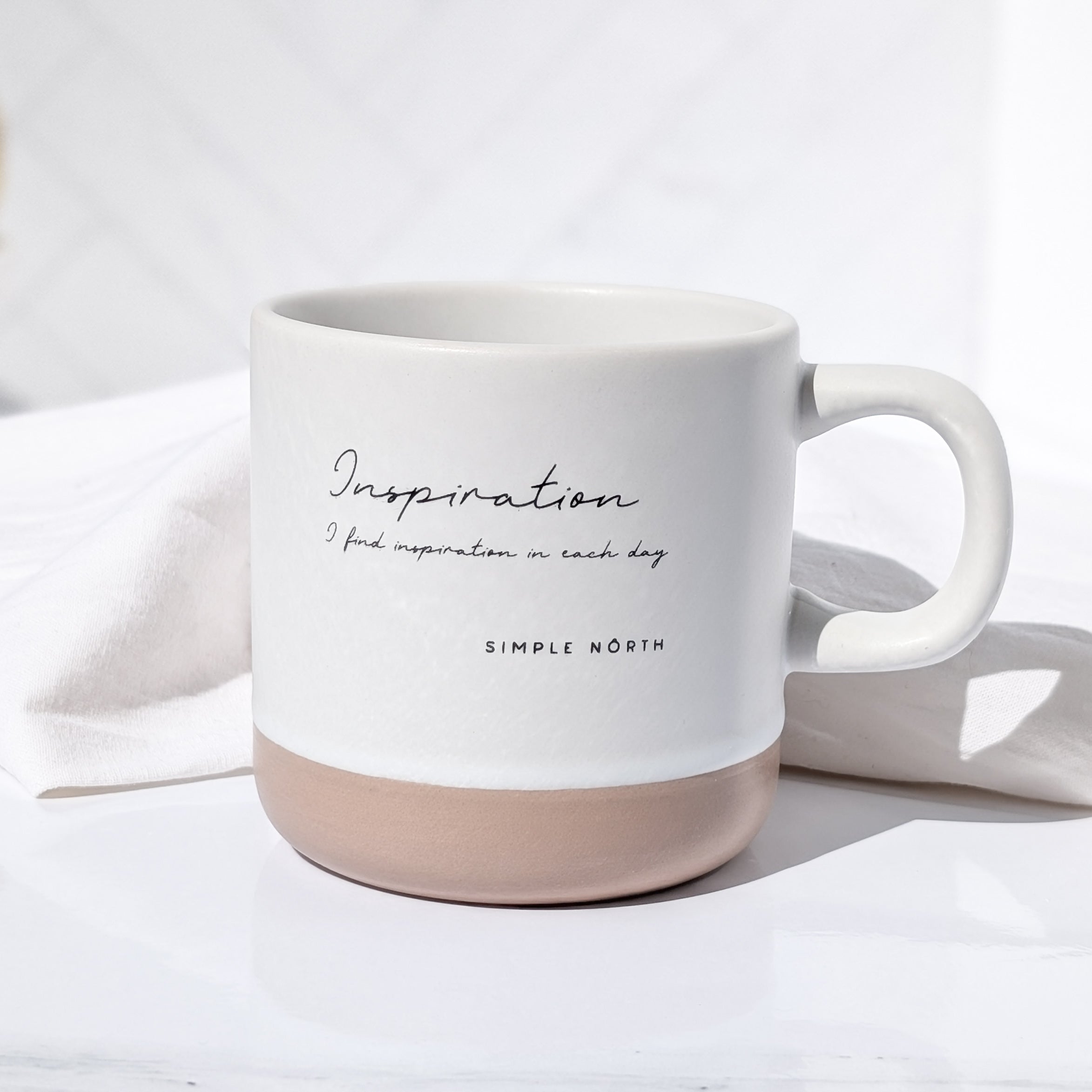 A white ceramic mug with 'Inspiration' written on the side along with a quote 'Simple North'. The mug has a natural clay base, and it is placed on a white surface with a light pink hue.